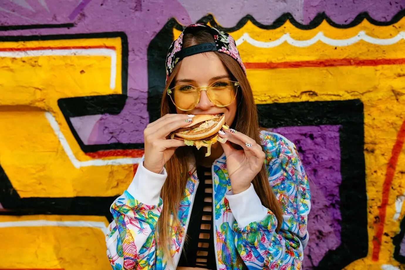 An attractive young woman eating a delicious burger, wearing a colorful jacket and a cap.